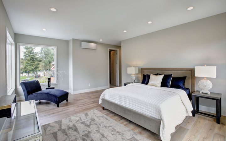 A well-lit bedroom with a large window offering a view of the outdoors. The room features a neatly made bed with white bedding and navy blue pillows, a navy blue armchair, and two bedside tables with lamps. The walls are painted a soft gray, and there is a patterned rug on the floor.