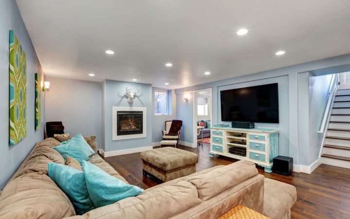 A comfortable living room with a beige sectional sofa adorned with blue throw pillows. The room features a fireplace, a mounted flat-screen TV, and a wooden entertainment unit. The walls are painted a soft blue, and there is a staircase leading to another level.