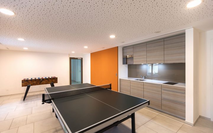 A spacious game room featuring a ping pong table, a foosball table, and a modern kitchenette with light wood cabinets. The room has a bright orange accent wall and recessed lighting in the ceiling.