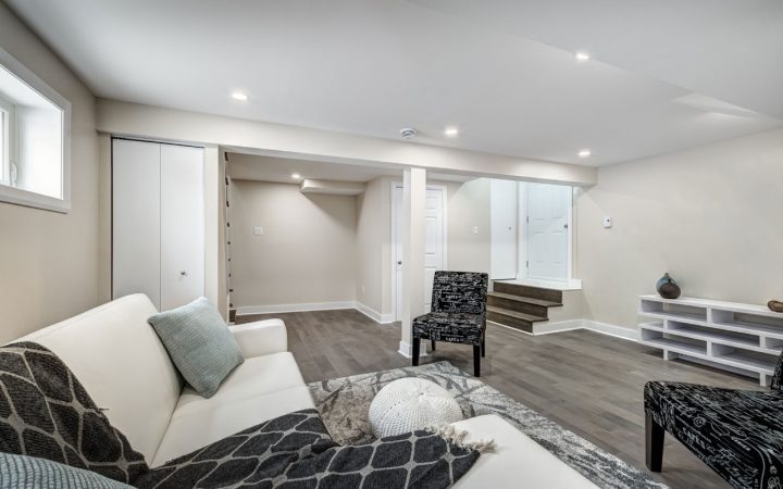 A cozy basement living area with a white sofa, a patterned armchair, and a small ottoman. The room has a modern design with recessed lighting and a staircase leading to another level. A white entertainment unit is placed against the wall.