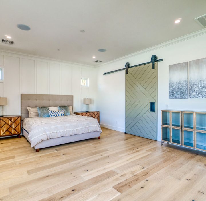 A stylish master bedroom with a large bed, two nightstands, and a sliding barn door. The room has hardwood flooring, white walls, and a built-in storage unit with glass doors. Decorative artwork hangs above the bed.