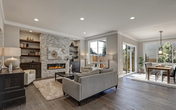 A warm and inviting living room with a stone fireplace as the focal point. The room features a gray sofa, a white armchair, and a wooden coffee table. Large windows let in natural light, and there is a dining area visible in the background.