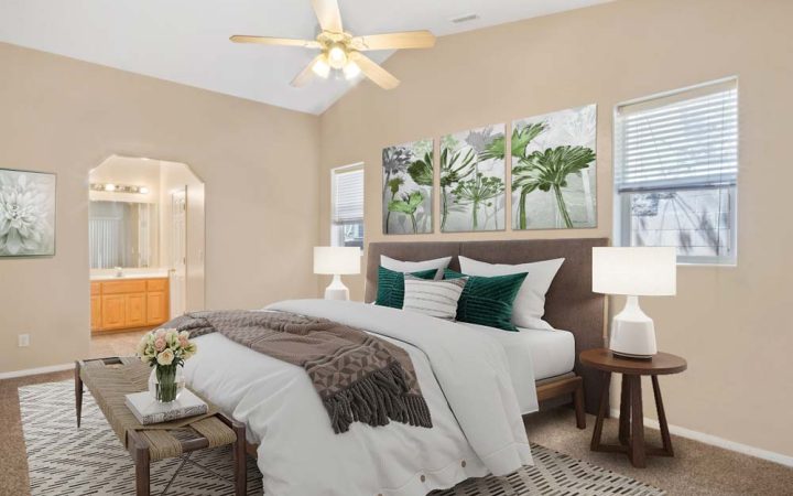 A serene bedroom with beige walls, a large bed adorned with green and white pillows, and two bedside tables with lamps. The room features a ceiling fan, a window with blinds, and a bench at the foot of the bed. There is also an en-suite bathroom visible through an archway.