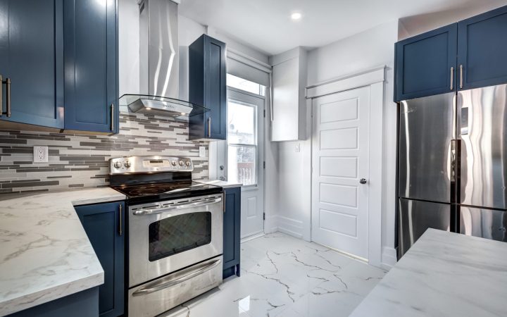 A contemporary kitchen featuring blue cabinets, a stainless steel oven, and a marble countertop. The backsplash is made of mosaic tiles, and the floor is covered in white marble tiles. A glass door leads to an outdoor area.